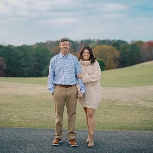 Cassidy-1_optim A couple standing on a paved path with grassy hills and trees in the background, the woman holding the man’s arm while wearing a beige knit dress and ankle boots.