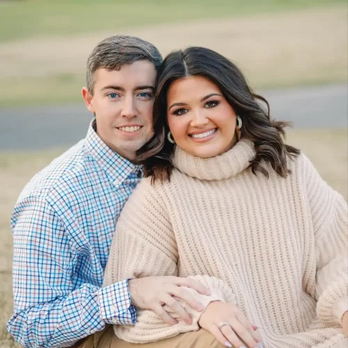 Cassidy 2 A smiling couple seated on dry grass in an open field, with the woman wearing a beige knit sweater dress and the man in a plaid button-down shirt and khaki pants.
