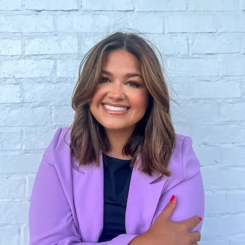 Cassidy Headshot Professional headshot of a smiling woman with shoulder-length brown hair, wearing a lavender blazer over a black top, posed against a light blue brick wall.