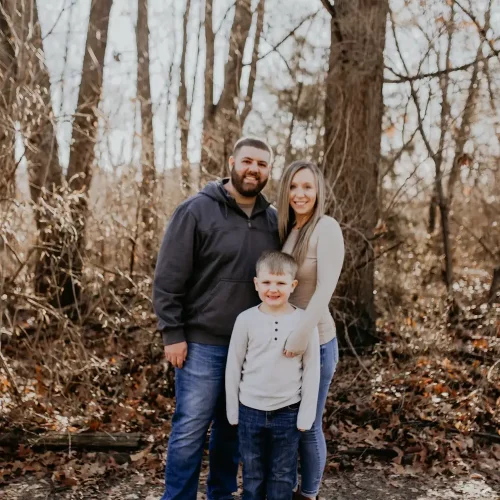 Kristina 1 A smiling family of three standing in a wooded area during autumn, with bare trees and fallen leaves on the ground. The parents stand behind a young boy in a light sweater and jeans.