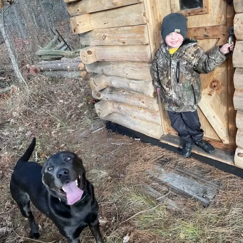 Kristina-4_q50 A young boy in a camouflage jacket and knit hat smiling at the entrance of a rustic log cabin, with a black dog in the foreground looking excited with its tongue out.