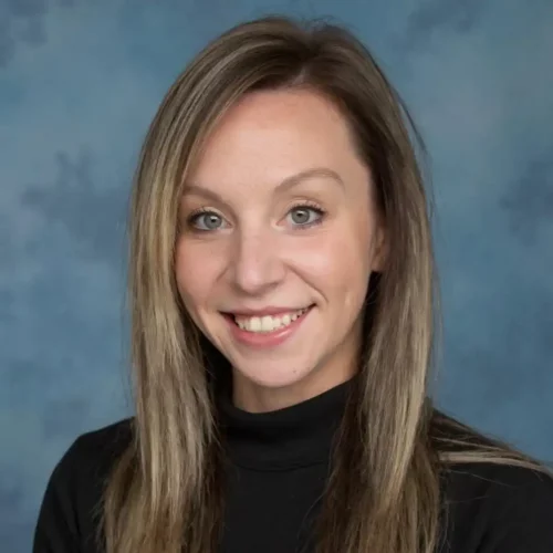 Kristina-5 Professional headshot of a smiling woman with straight, highlighted hair wearing a black turtleneck, posed in front of a soft blue studio background.
